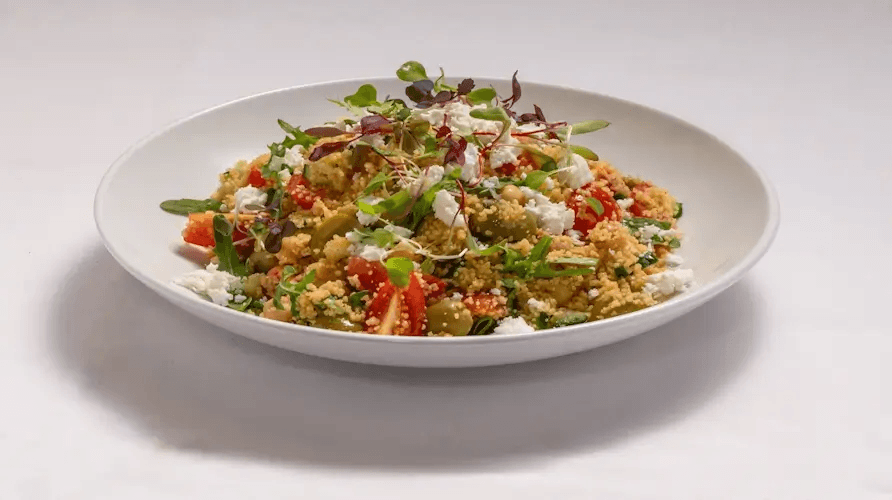 White bowl filled with a colorful grain salad—crumbled cheese, bright tomatoes, greens, and microgreens on a white plate, ready to serve.
