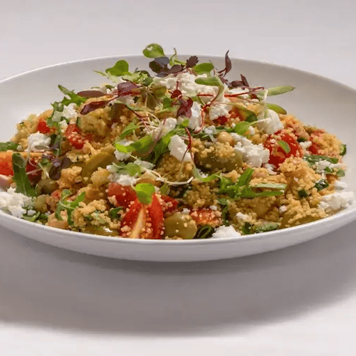 White bowl filled with a colorful grain salad—crumbled cheese, bright tomatoes, greens, and microgreens on a white plate, ready to serve.